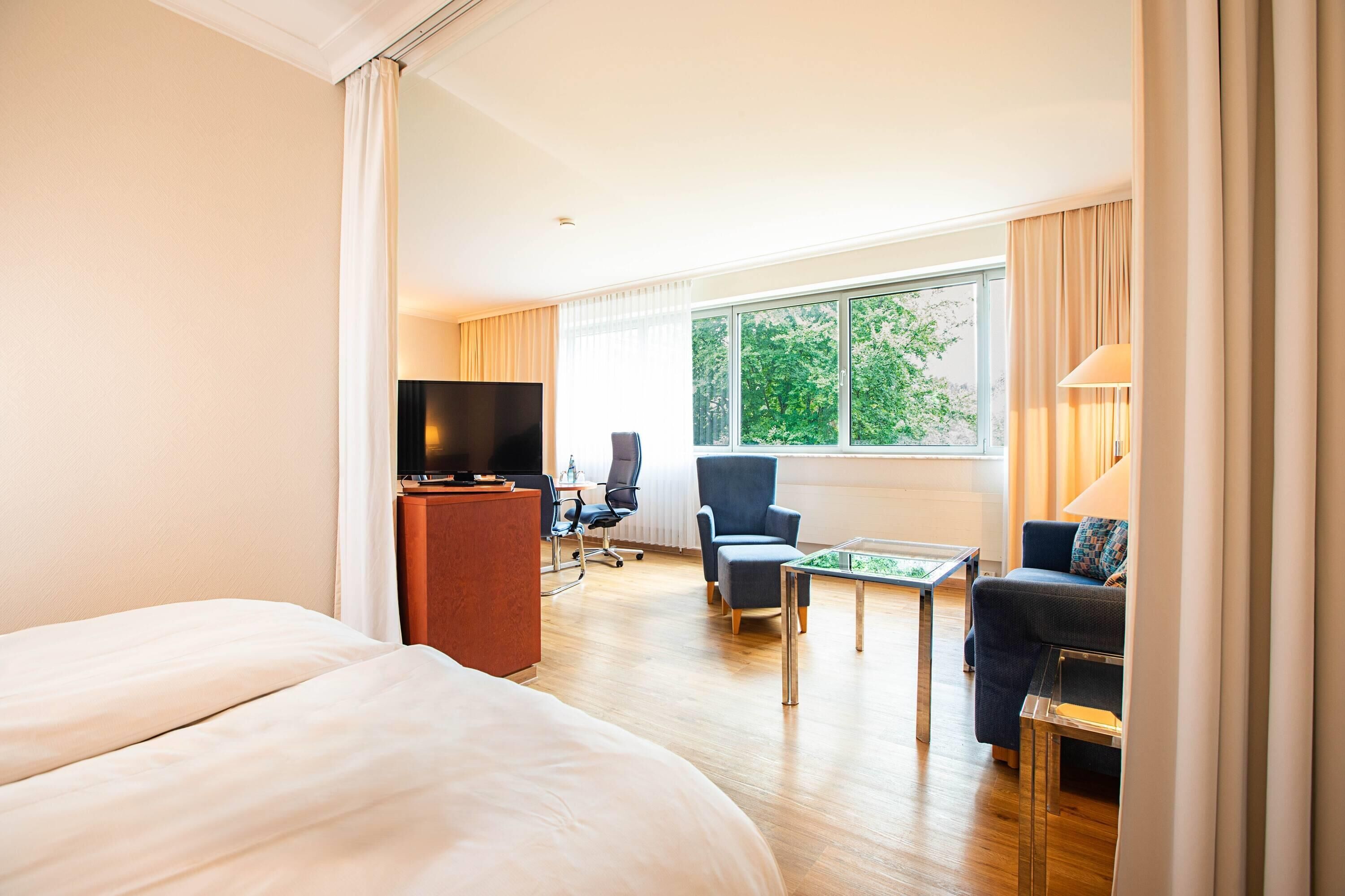 A hotel room with a bed, a desk, a television, a chair, a sofa, and a window with a view of trees.
