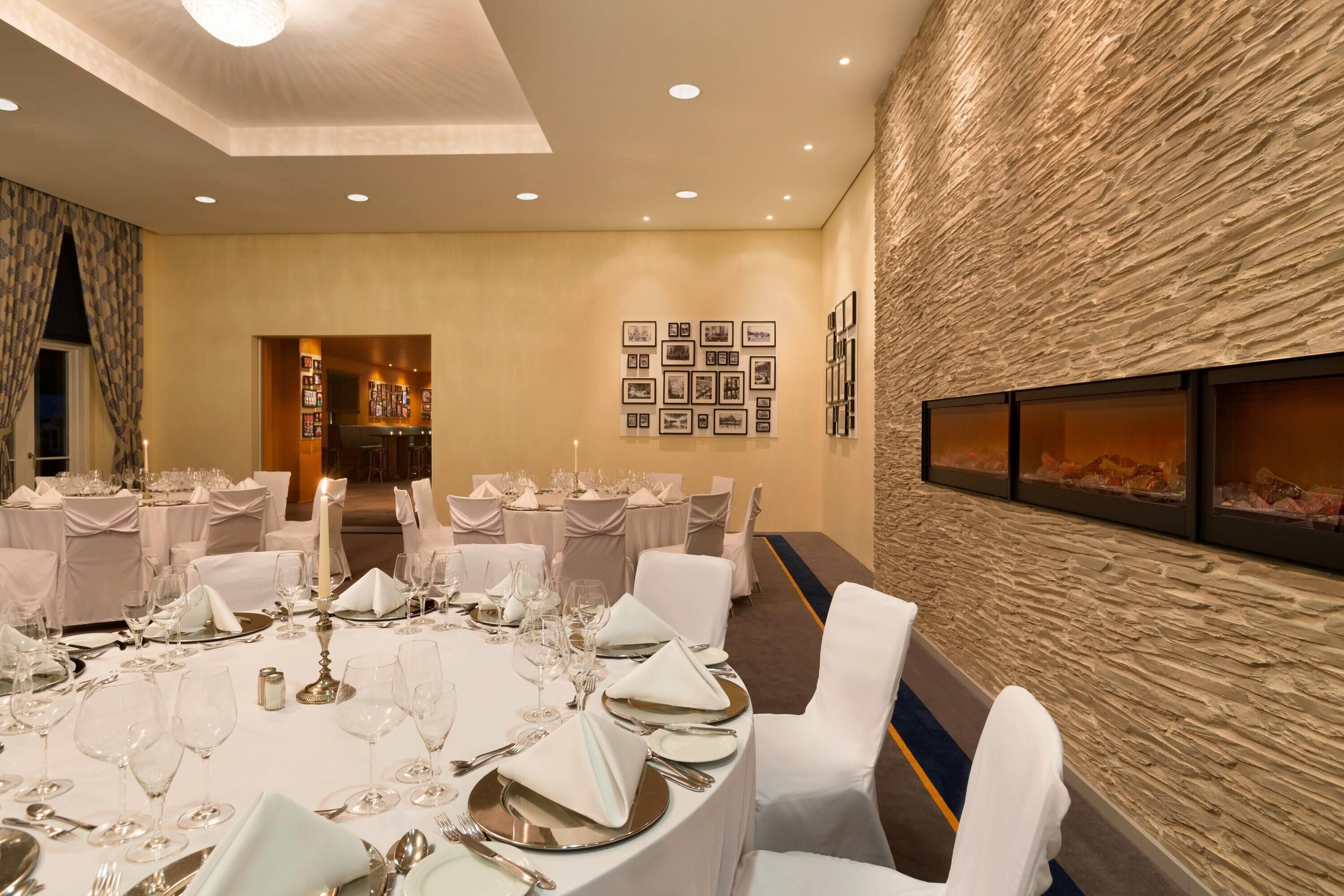 Un salón de banquetes con mesas redondas dispuestas para comer, una pared de piedra con cuadros enmarcados y una chimenea.
