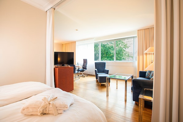 A hotel room with a bed, a desk, a television, a chair, a sofa, and a window with a view of trees.