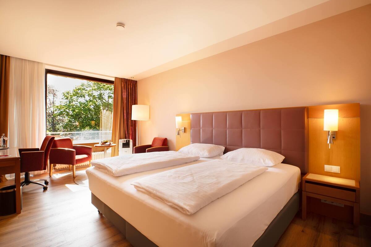 Une chambre d’hôtel avec deux lits, un bureau, une chaise et une fenêtre donnant sur des arbres.