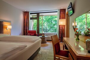 Habitación de hotel con cama, escritorio, silla, televisor y ventana con vista a árboles.