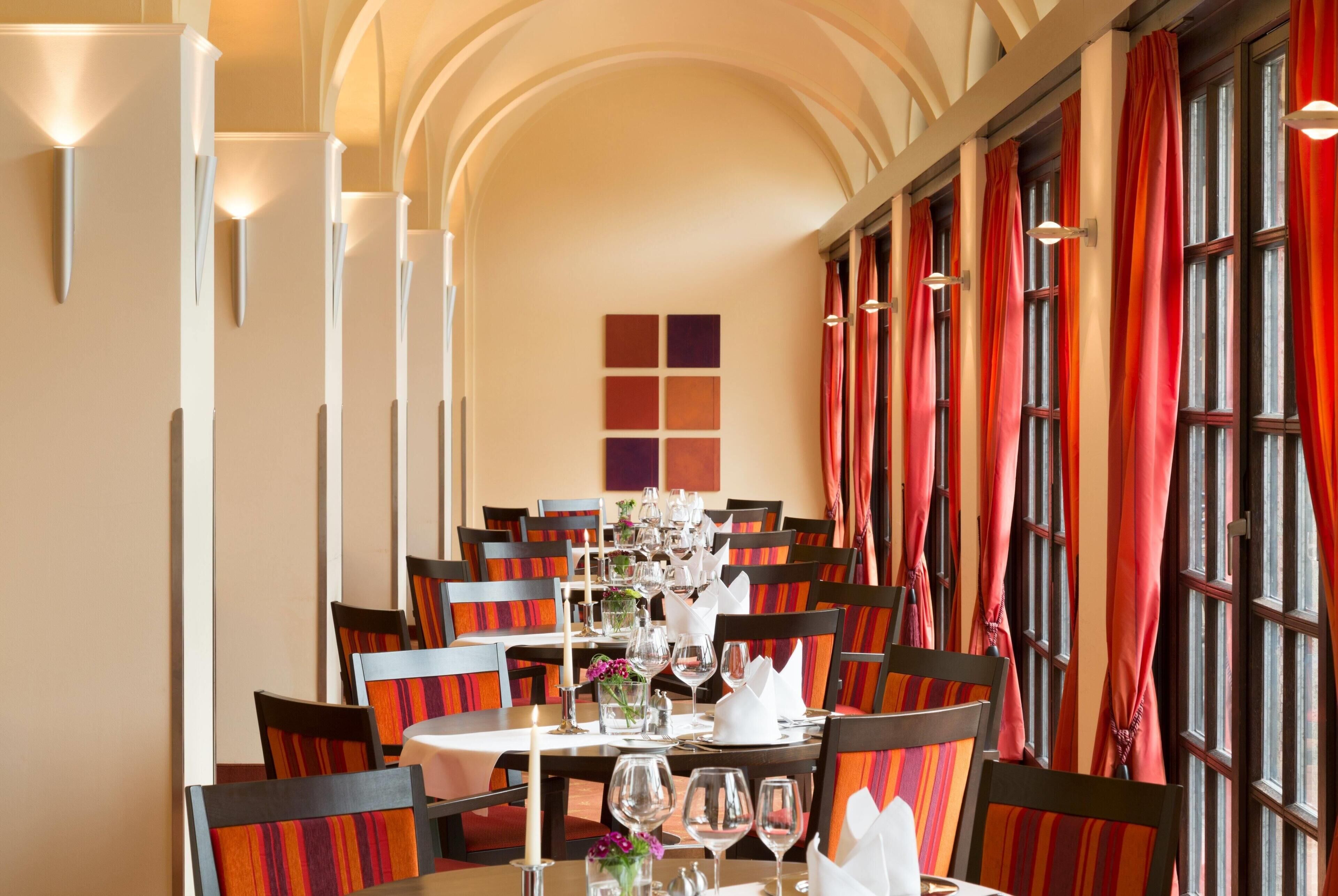 A dining area with tables set for service, chairs with striped upholstery, and large windows with red curtains.