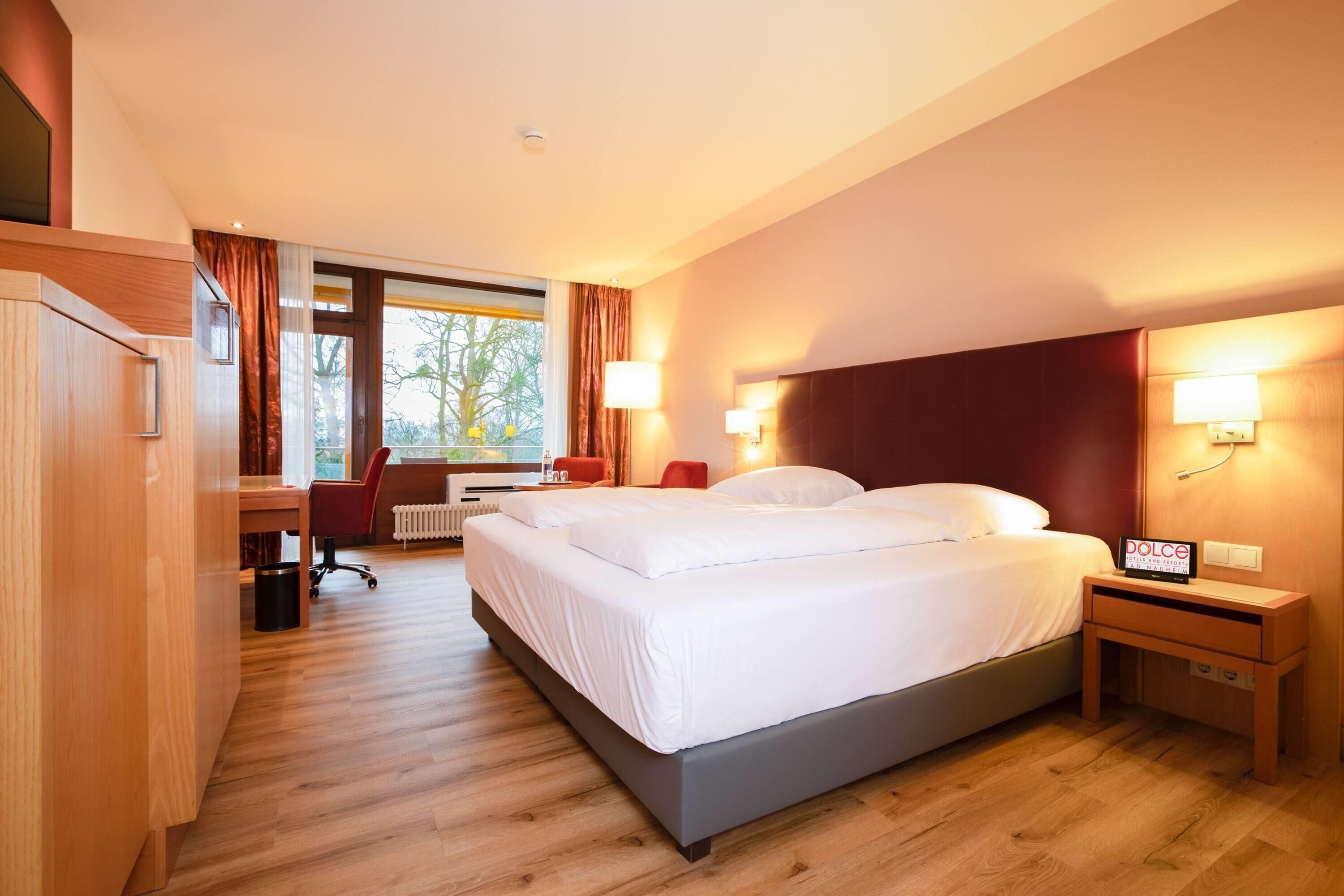 A hotel room with a bed, bedside table, desk, chair, and a view of the outdoors.