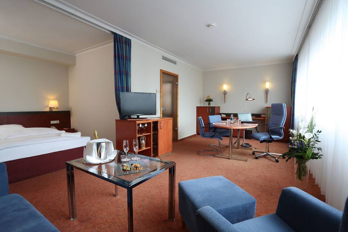 Une chambre d’hôtel comprenant un lit, une télévision, un bureau avec un ordinateur portable, deux chaises, une petite table avec un verre et un vase de fleurs.