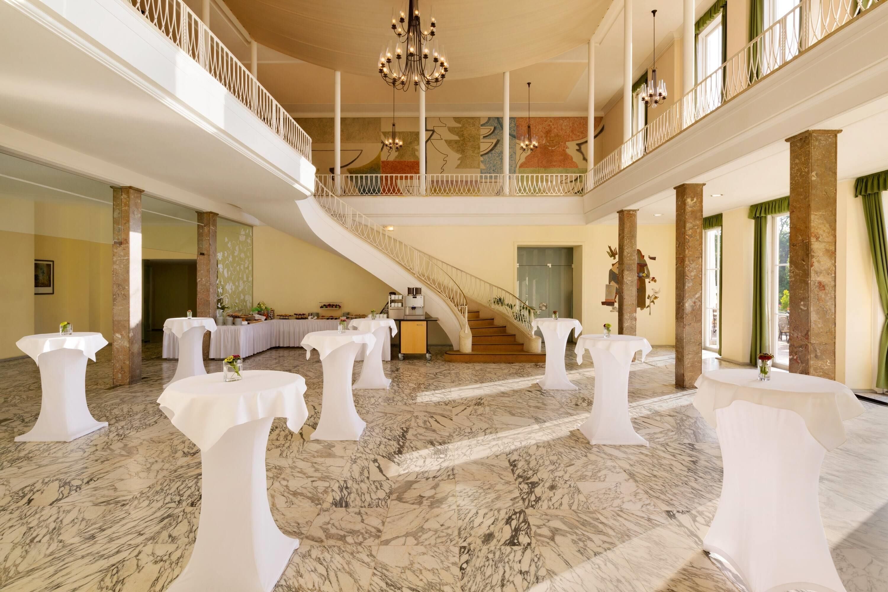 Un vaste hall doté d’un sol en marbre, de tables blanches, d’un magnifique escalier et d’un balcon agrémenté de balustrades.