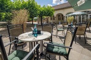 Eine Außenpatio mit Korbstühlen und Tischen, einer Wasserflasche auf einem Tisch und einem Gebäude mit Bögen im Hintergrund.