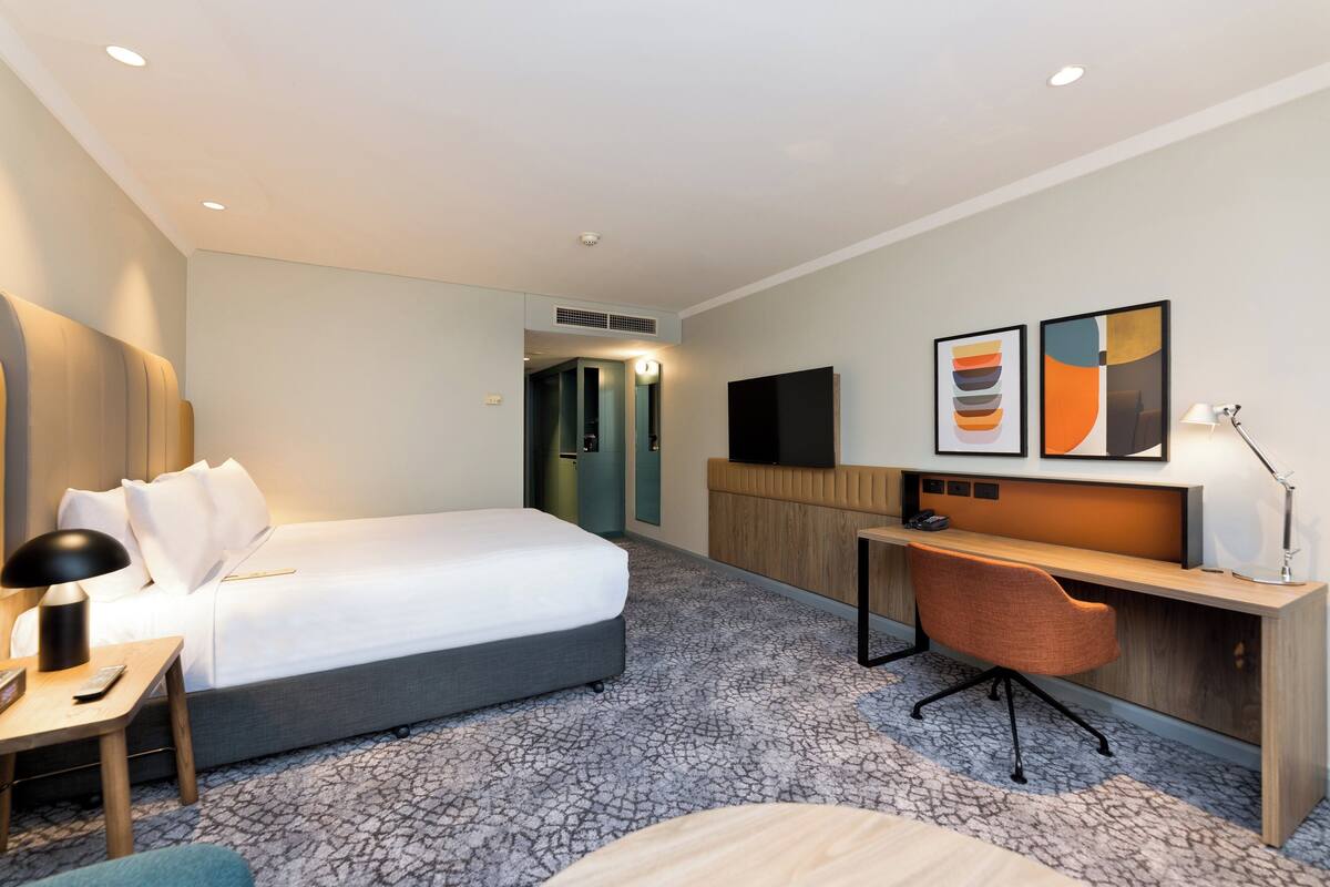 A hotel room with a bed, desk, chair, and artwork on the wall.
