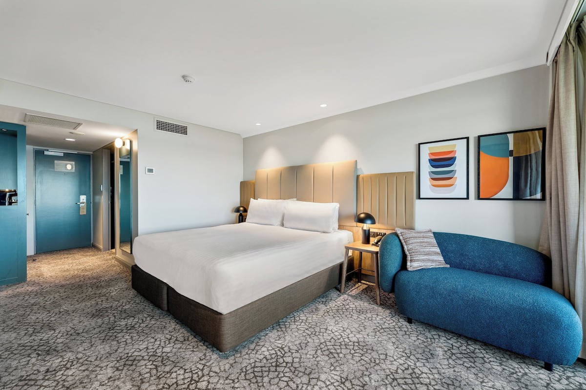 A hotel room with a large bed, a desk, a blue sofa, and two framed artworks on the wall.