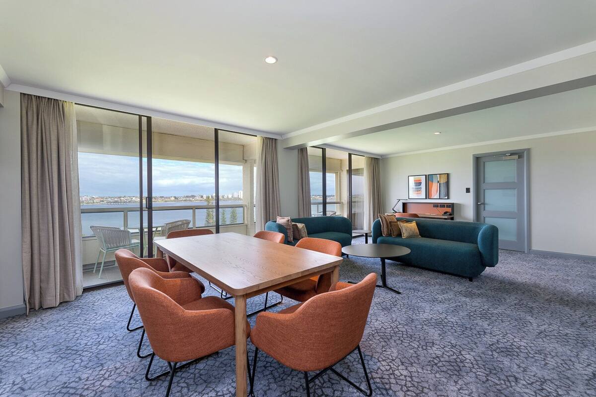 A hotel room with a large window offering a city view, a dining table with chairs, a sofa, and a small desk with a lamp.