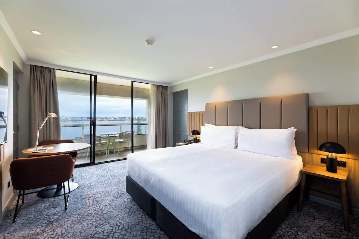 A hotel room with a large bed, a desk with a chair, a lamp, and a view of the city through a large window.