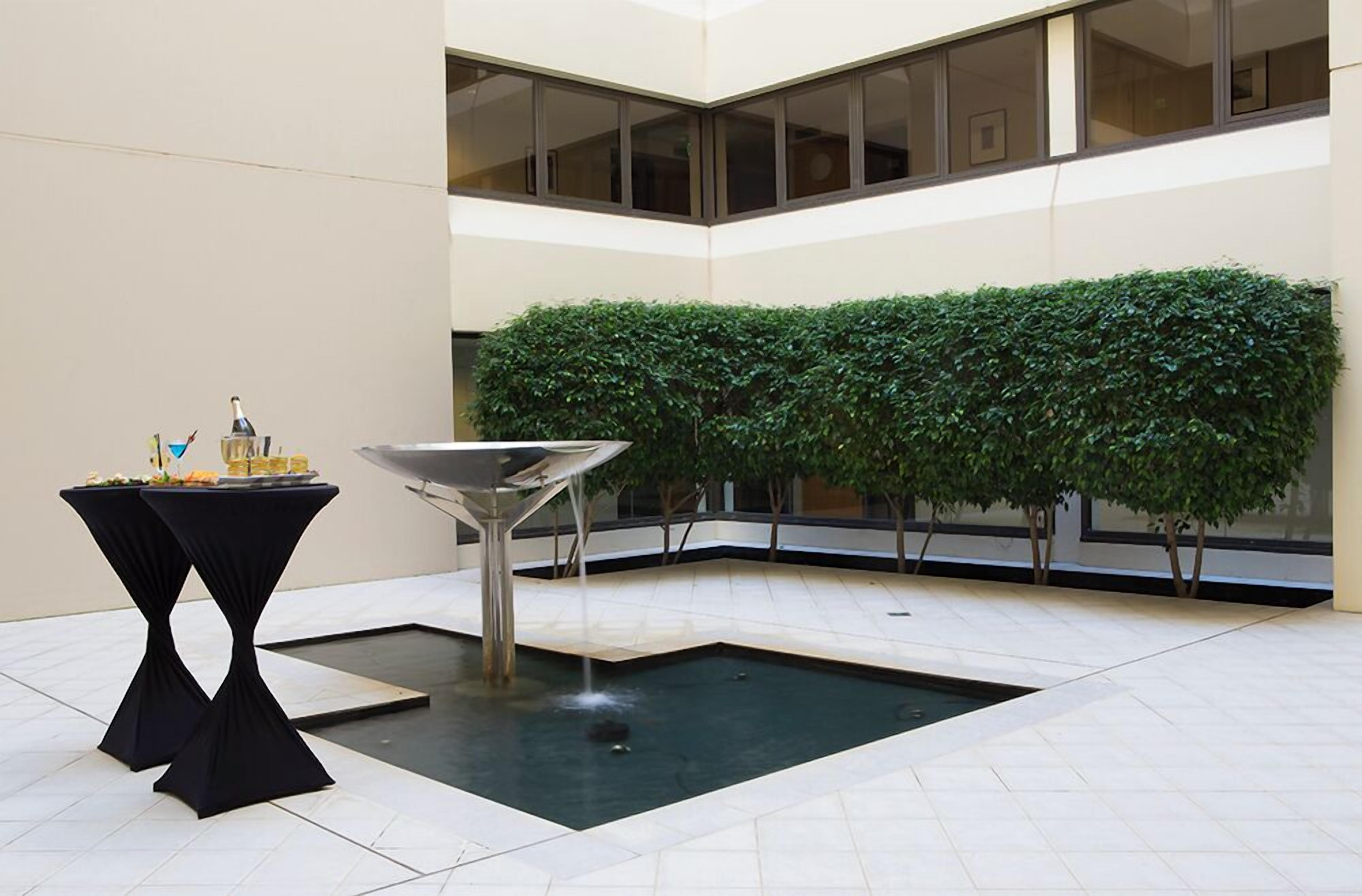 Un patio interior moderno con una fuente central, una mesa negra con un adorno y un seto recortado.