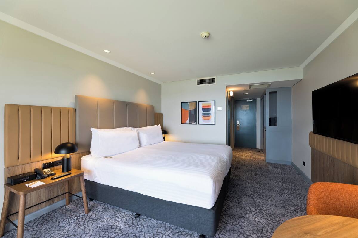 A modern hotel room with a large bed, a bedside table with a lamp, a flat-screen TV, and a small seating area.