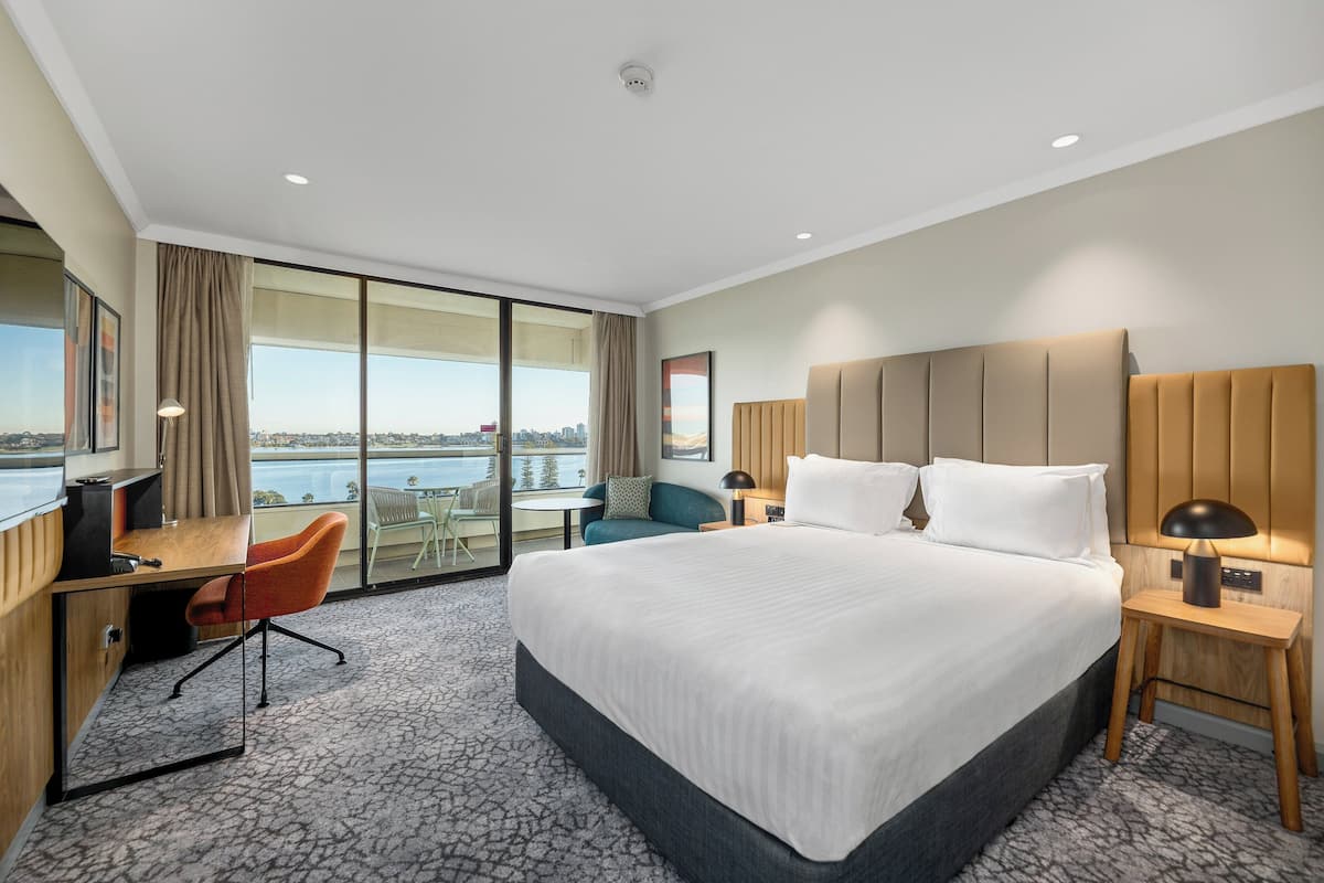 A hotel room with a large bed, a desk, a chair, a lamp, and a view of the city.