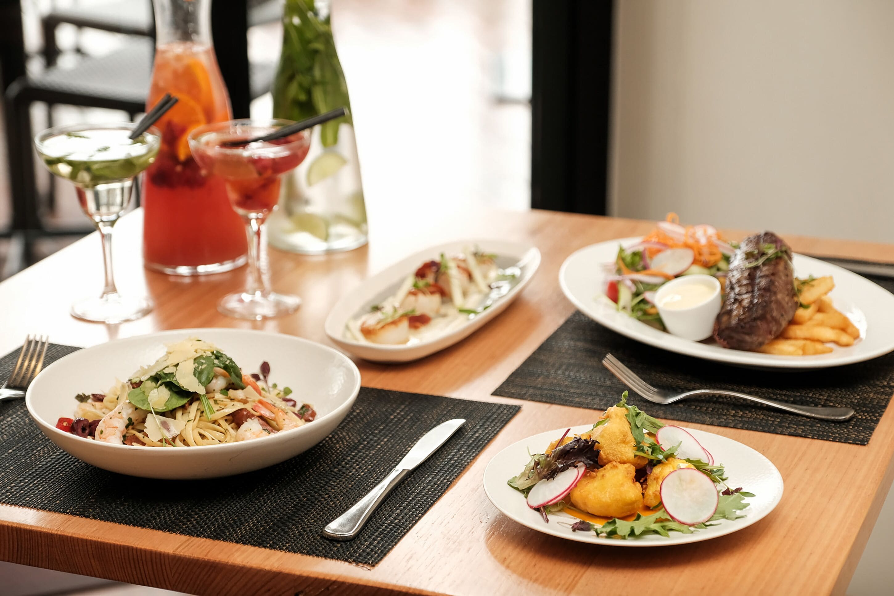 A dining table with various dishes, including a salad, a plate of fries, and a steak, along with two cocktails.
