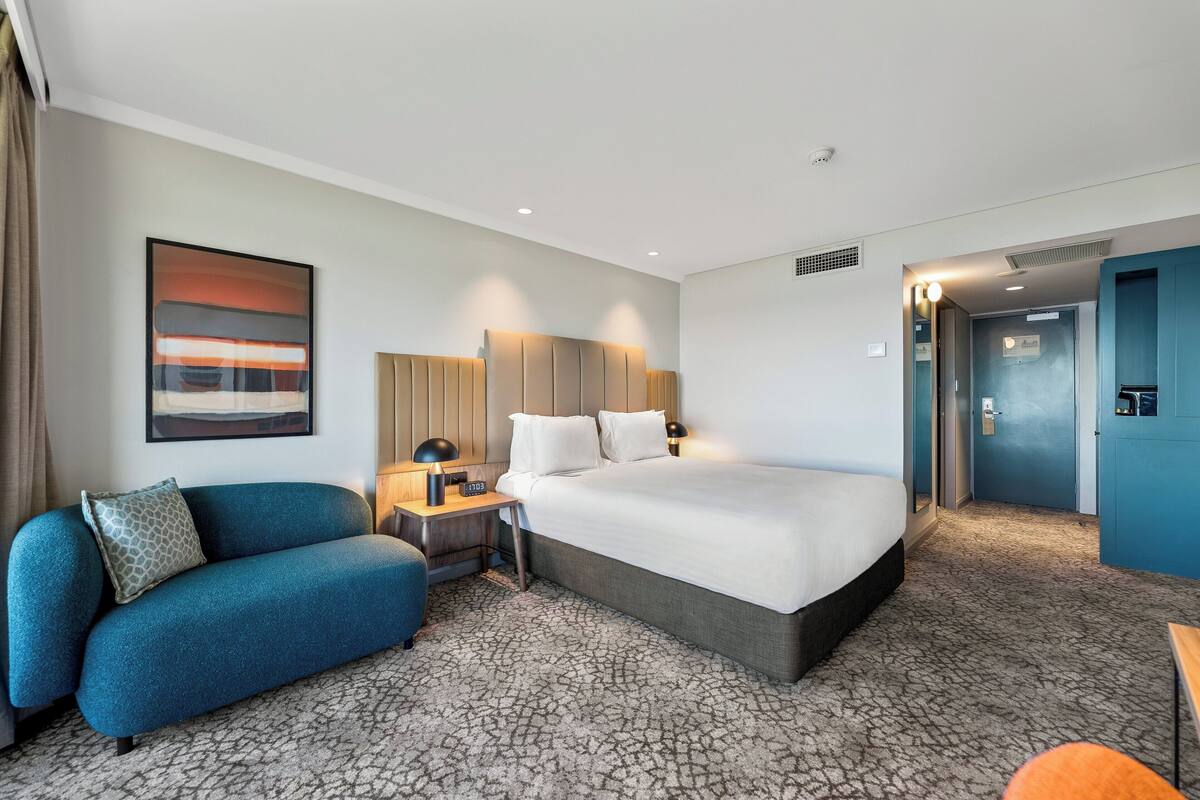 A hotel room with a large bed, a sofa, a desk with a lamp, and a painting on the wall.