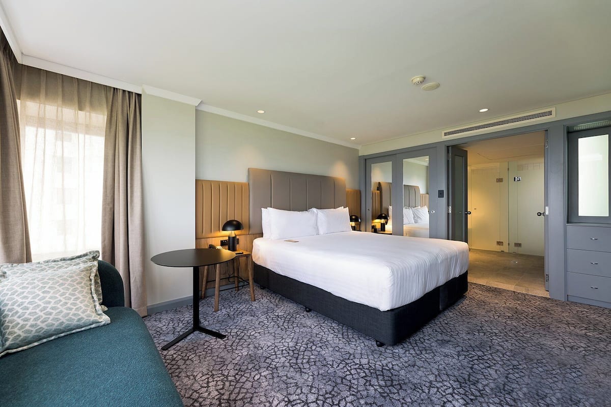 A hotel room with a large bed, a bedside table, a desk, and a chair. There is a window with curtains, a door, and a mirror.