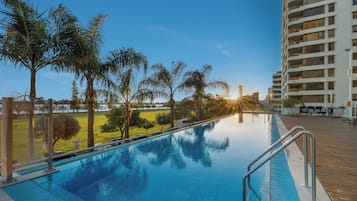 A swimming pool with a wooden deck, surrounded by palm trees and a high-rise building in the background.