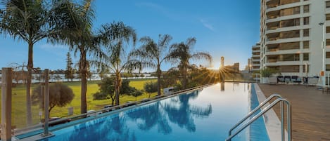A swimming pool with a wooden deck, surrounded by palm trees and a high-rise building in the background.