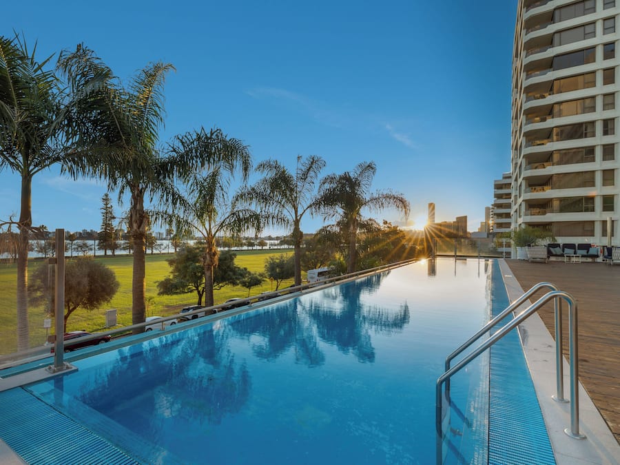 A swimming pool with a wooden deck, surrounded by palm trees and a high-rise building in the background.