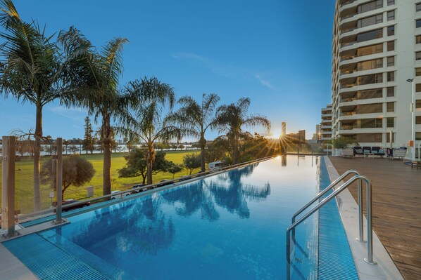 A swimming pool with a wooden deck, surrounded by palm trees and a high-rise building in the background.