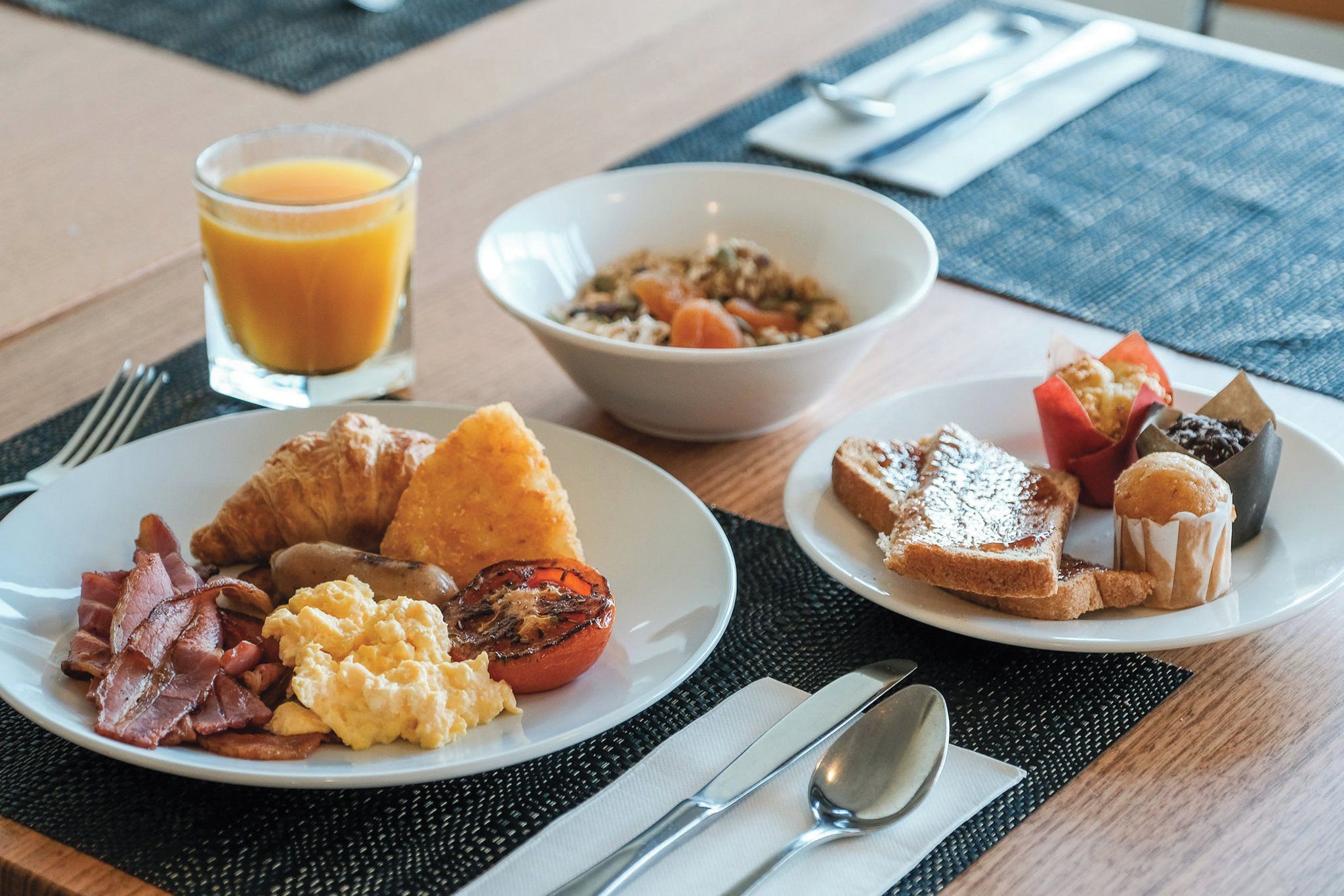Una mesa de desayuno con platos de huevos, tocino, tostadas y pasteles, acompañada de un vaso de jugo de naranja y un tazón de cereal.