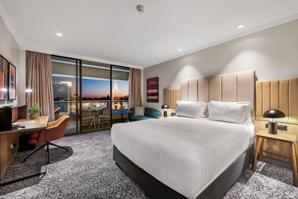 A modern hotel room with a large bed, a desk, a chair, and a view of the cityscape.