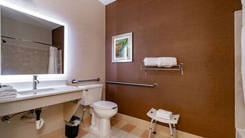 Une salle de bain avec un lavabo blanc, des toilettes et des barres d’appui. Un tableau encadré est accroché au mur, et une petite table avec une serviette s’y trouve.