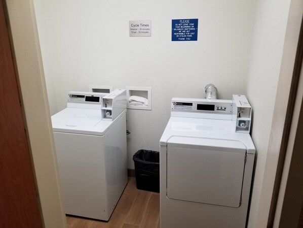A laundry area with two white washing machines, a sign indicating cycle times, and a trash bin. - Wingate by Wyndham Great Falls (Great Falls)