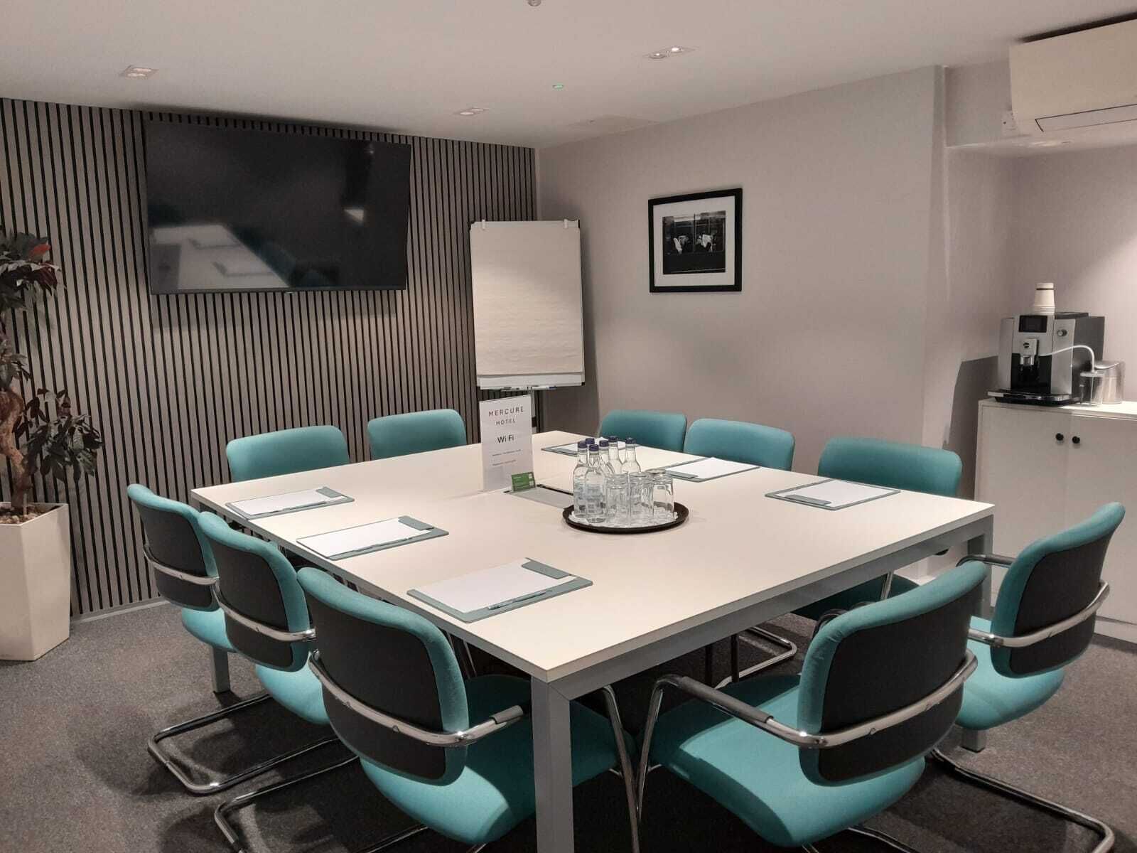 Ein Konferenzraum mit einem weißen Tisch, petrolfarbenen Stühlen, einem Wand-Fernseher, einer Whiteboard-Tafel und einer Kaffeemaschine.