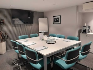 A conference room with a white table, teal chairs, a mounted TV, a whiteboard, and a coffee machine. - Mercure London Paddington (London)
