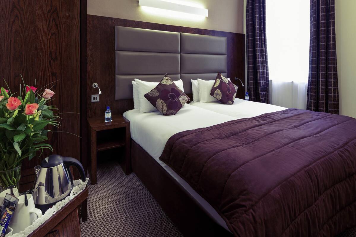 A hotel room with a large bed, a bedside table with a water bottle, a kettle, and a tray with cups and a teapot.