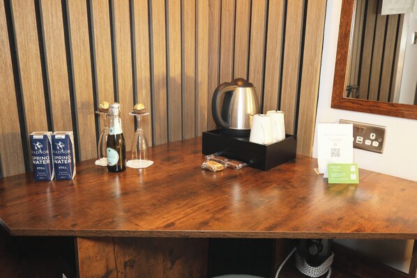 A wooden table with a bottle of Still, a glass, a box of pastries, a kettle, and a Mercure brochure. - Mercure London Paddington (London)