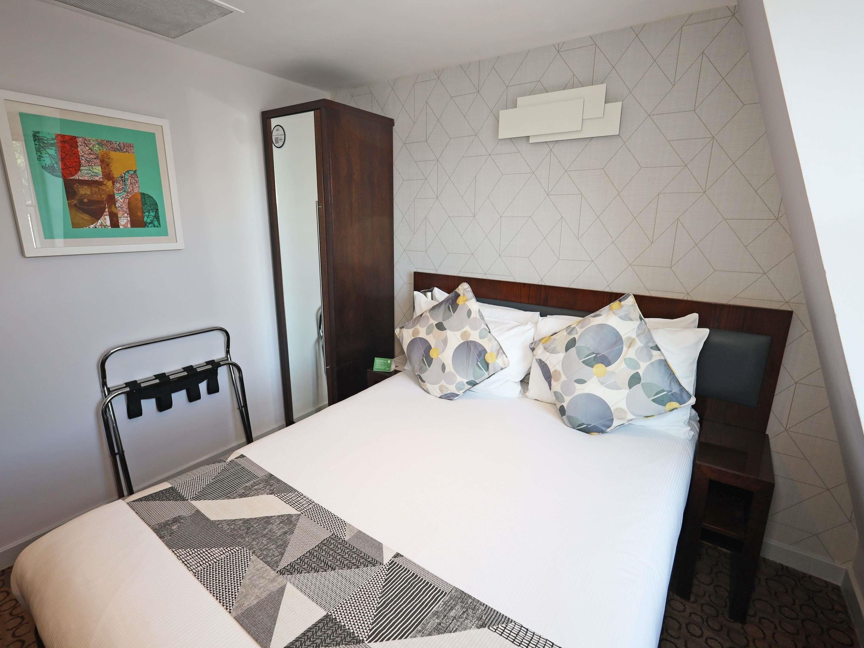 A hotel room with a bed, a bedside table, a chair, a coat rack, and a wall with geometric patterns.