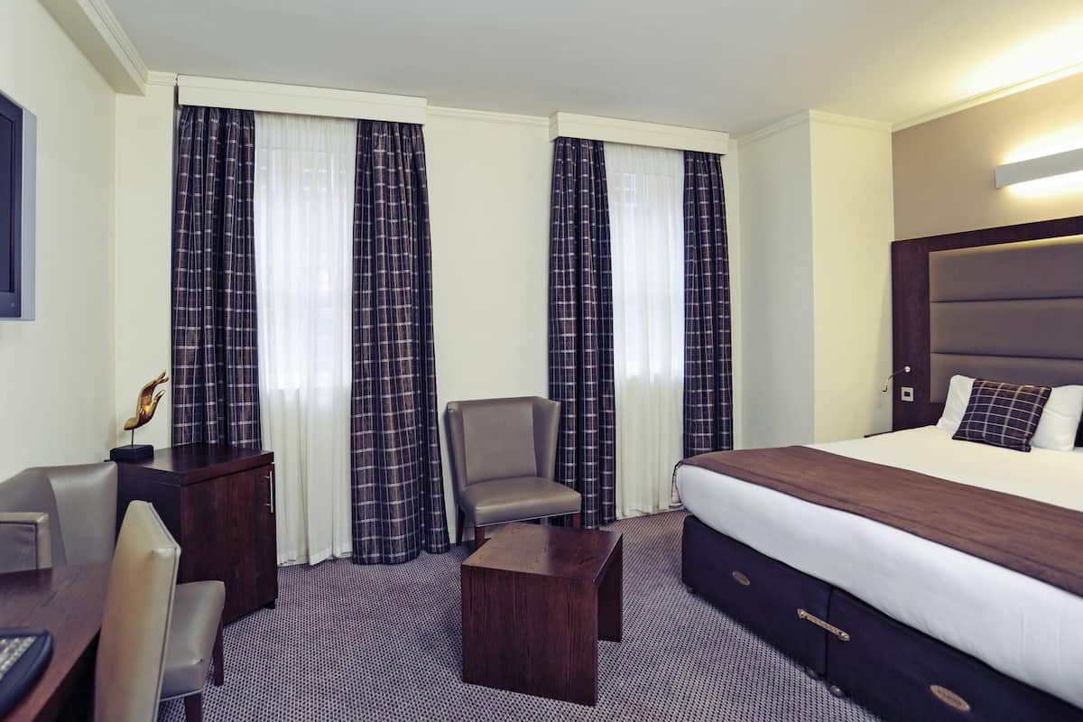 Une chambre d’hôtel avec un lit, un bureau, une chaise, une petite table et des rideaux.