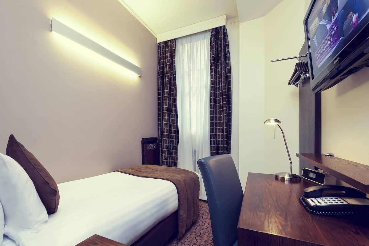 A hotel room with a bed, a desk with a telephone, a TV mounted on the wall, and a window with curtains.