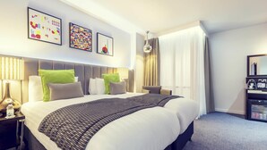 A hotel room with a large bed, a bedside table with a lamp, a minibar, and artwork on the wall. - Mercure London Paddington (London)