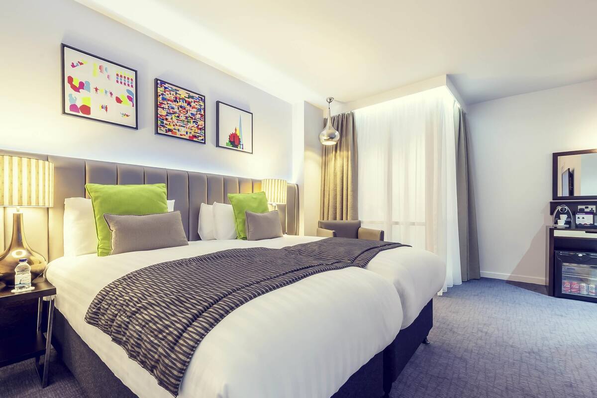 A hotel room with a large bed, a bedside table with a lamp, a minibar, and artwork on the wall.