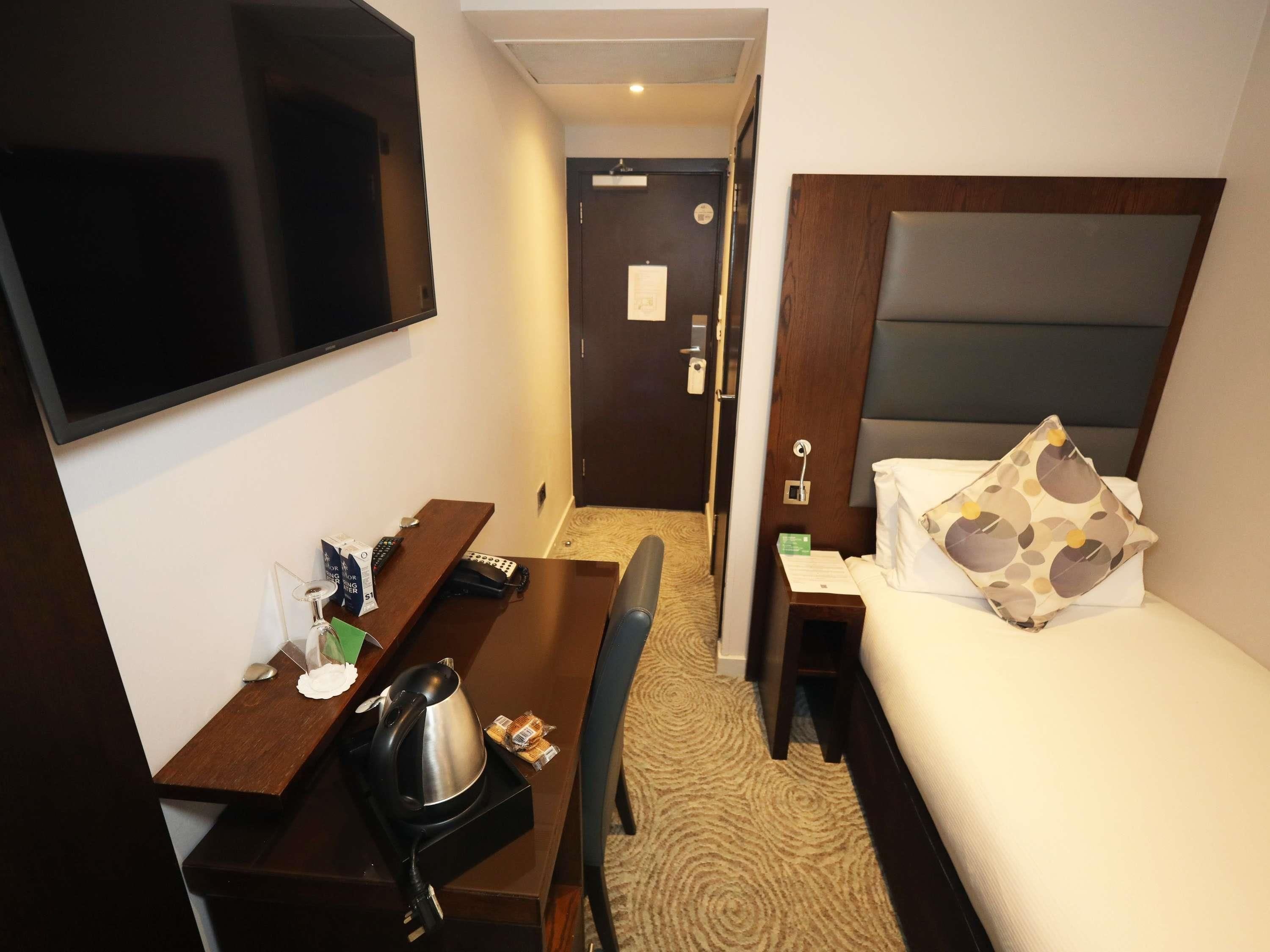 A hotel room with a bed, a desk with a kettle, a chair, a TV, and a door leading to a bathroom.