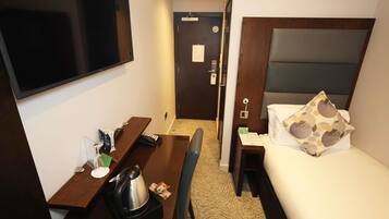 A hotel room with a bed, a desk with a kettle, a chair, a TV, and a door leading to a bathroom.