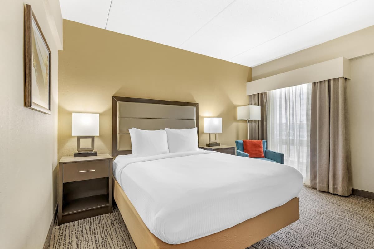 Une chambre d’hôtel avec un grand lit, deux tables de chevet avec des lampes, une chaise, une fenêtre avec des rideaux et un tableau encadré au mur.