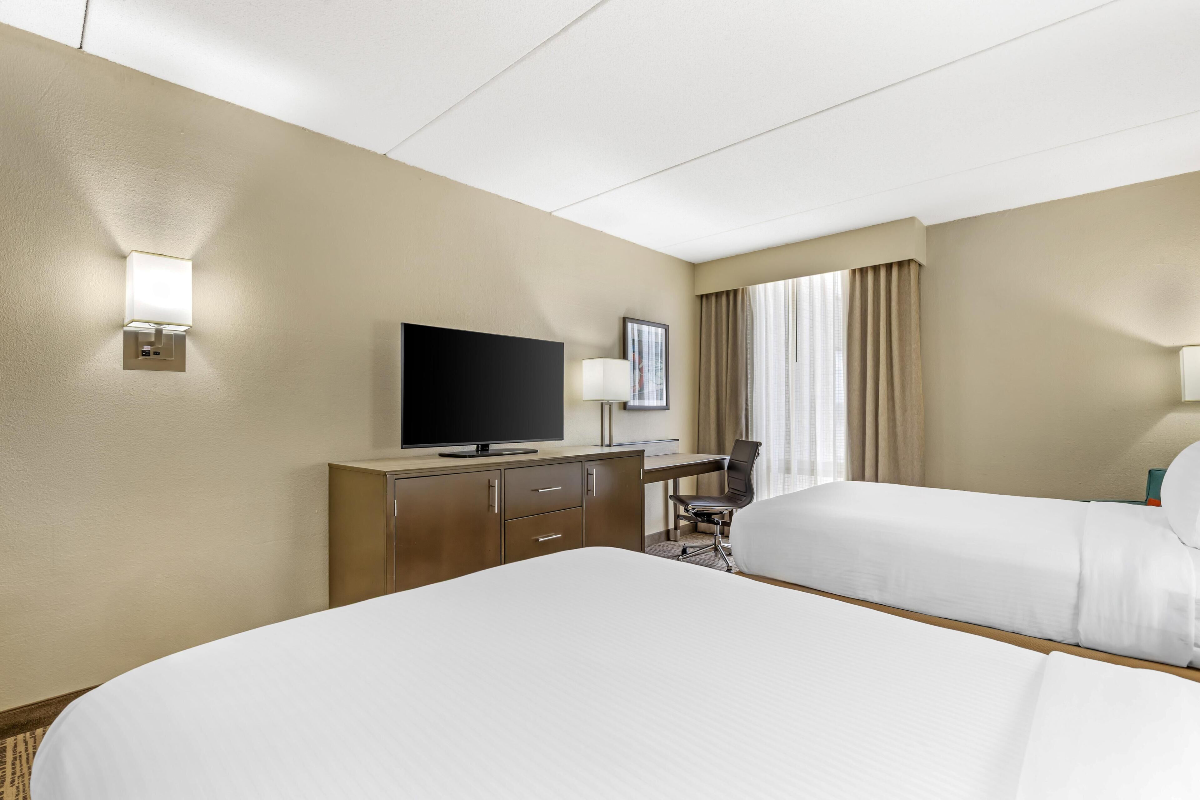 A hotel room with two beds, a desk with a flat-screen TV, a chair, and a window with curtains.