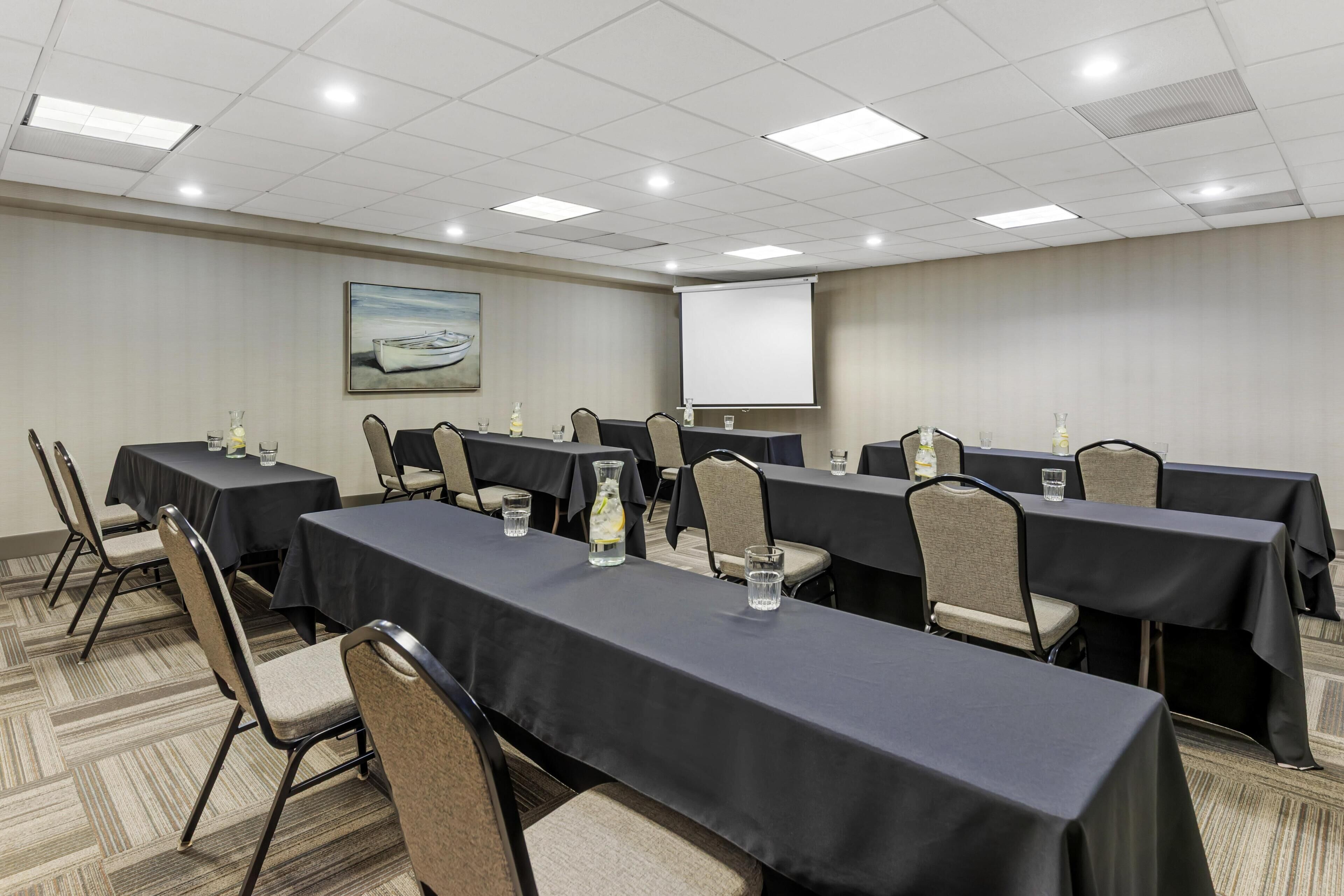 A conference room with multiple tables, chairs, a projector screen, and a painting on the wall.