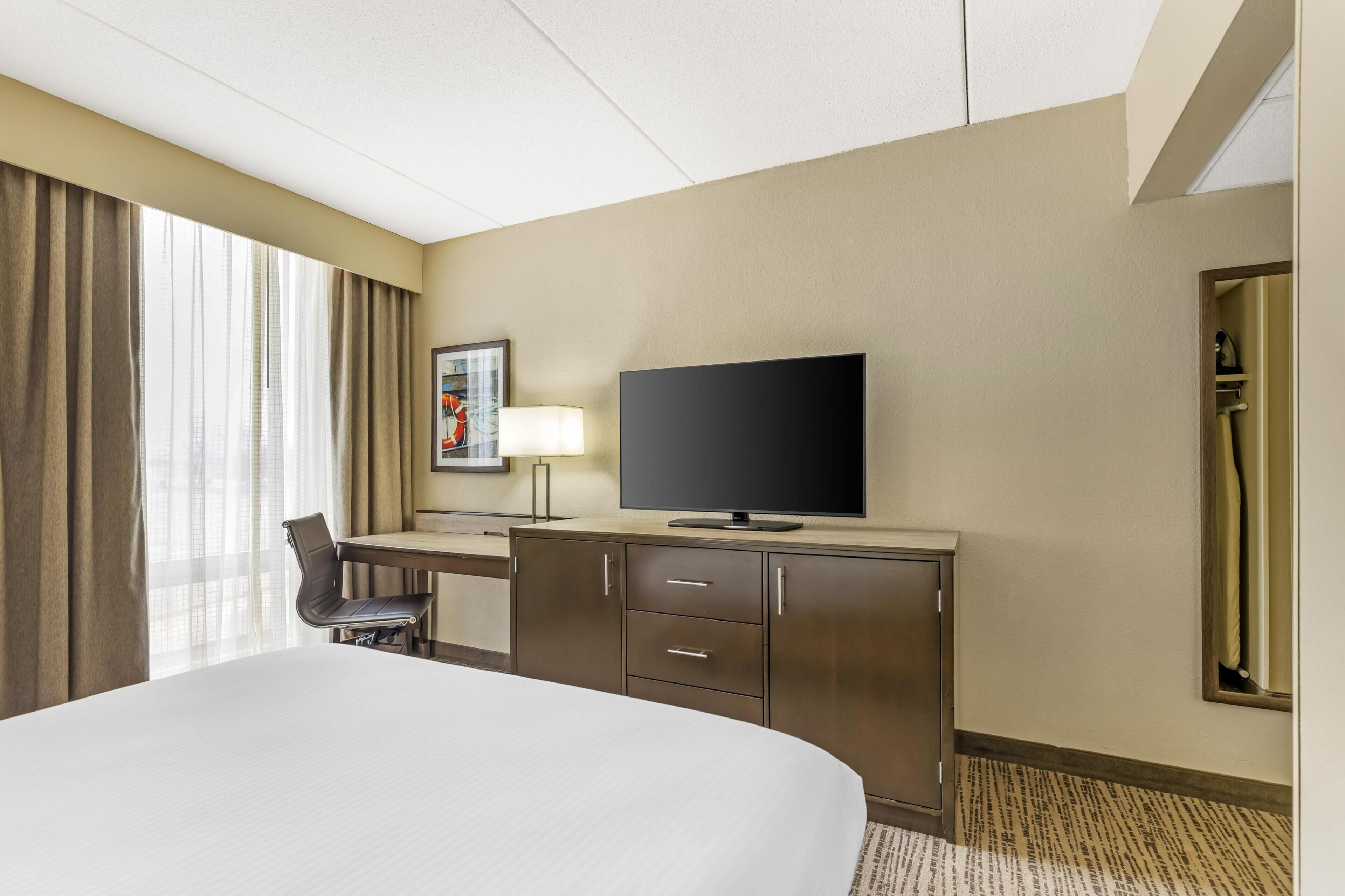 Habitación de hotel con cama, escritorio, televisión de pantalla plana, silla, lámpara de escritorio y ventana con cortinas.