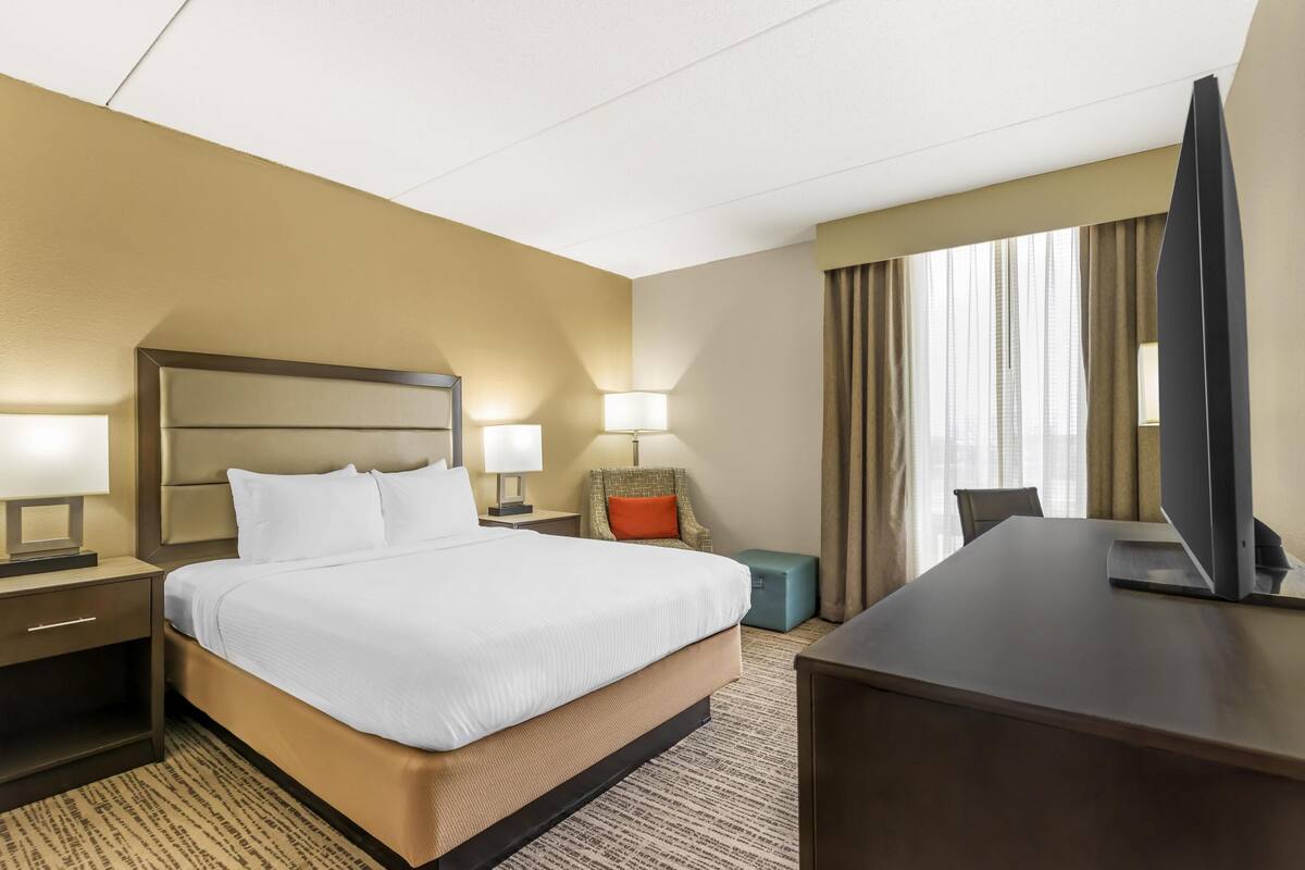 Une chambre d’hôtel comprenant un lit, des tables de chevet, un bureau, une télévision et une fenêtre avec des rideaux.