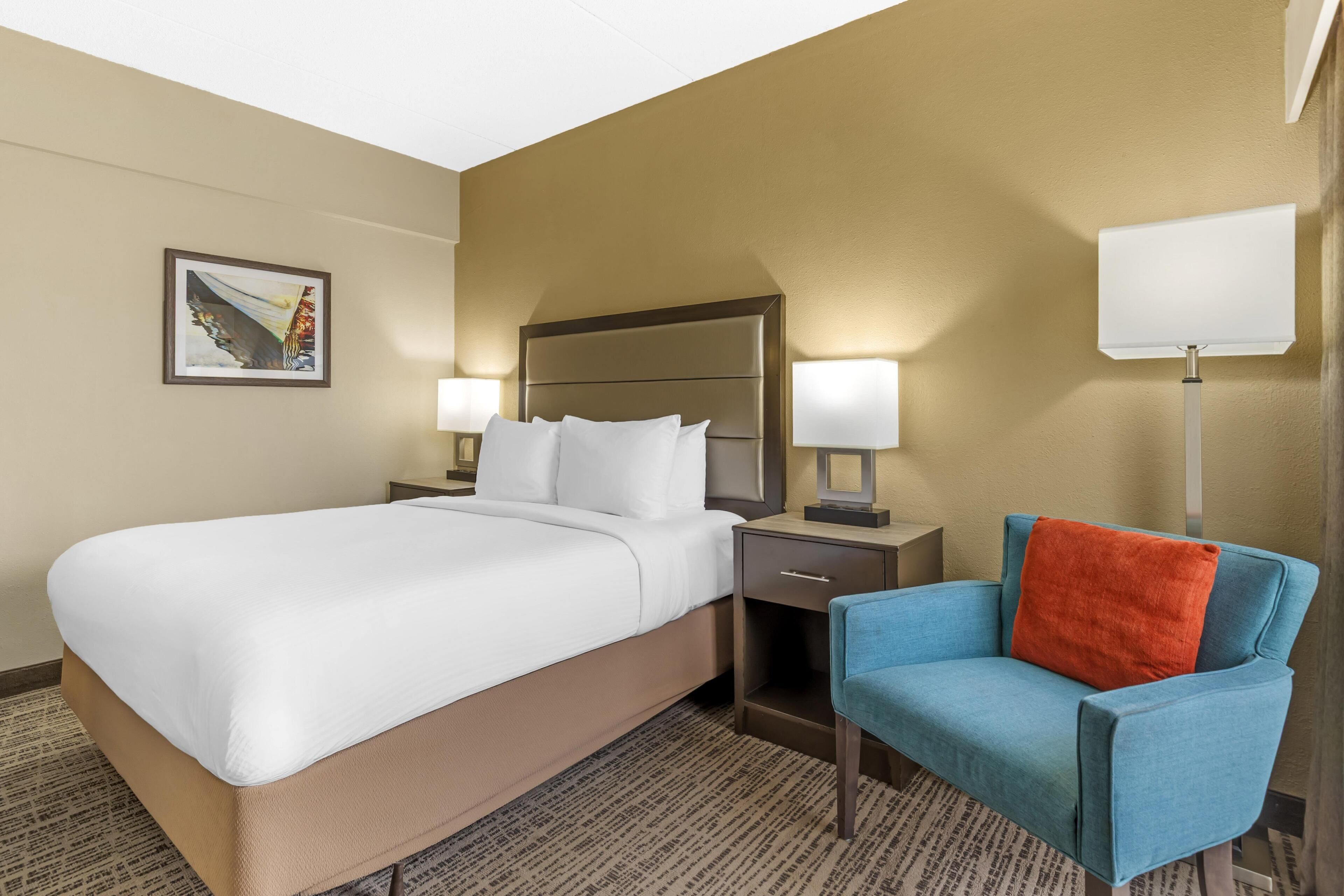 Une chambre d’hôtel avec un grand lit, deux tables de chevet avec des lampes, une chaise avec un coussin rouge et un tableau au mur.