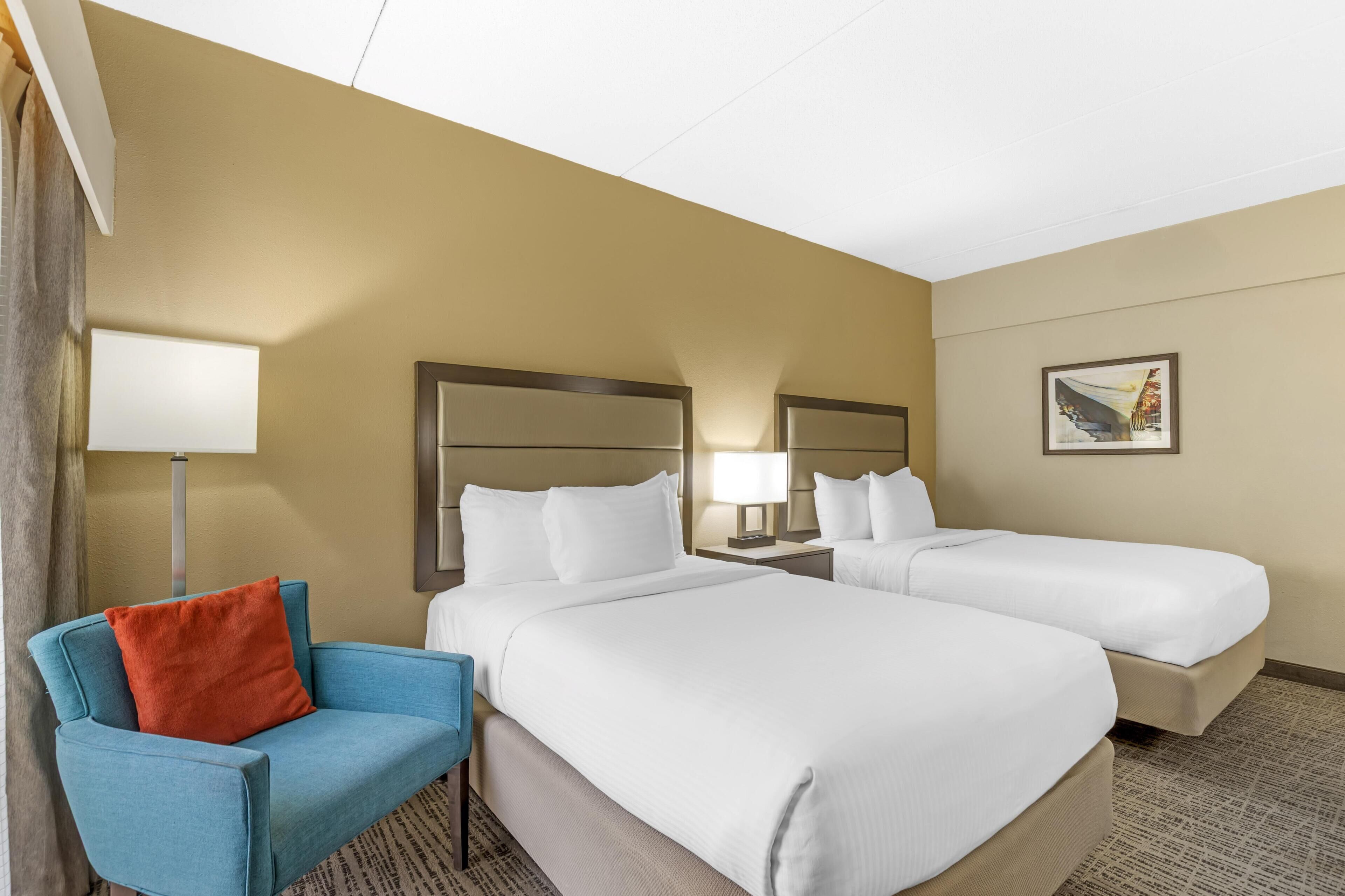 A hotel room with two beds, a blue chair, a lamp, and a painting on the wall.