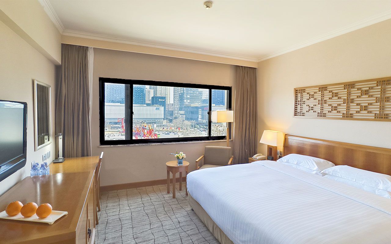 A hotel room with a large bed, a desk with a television, a small table with a vase, and a view of the city through large windows.