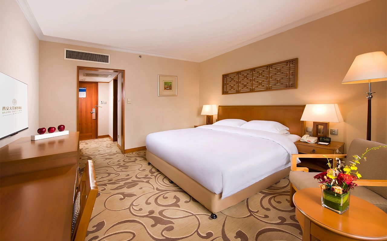 Une chambre d’hôtel avec un lit, des tables de chevet, un bureau et une chaise. Un élément décoratif orne le mur au-dessus du lit, et une petite table porte un vase rempli de fleurs.