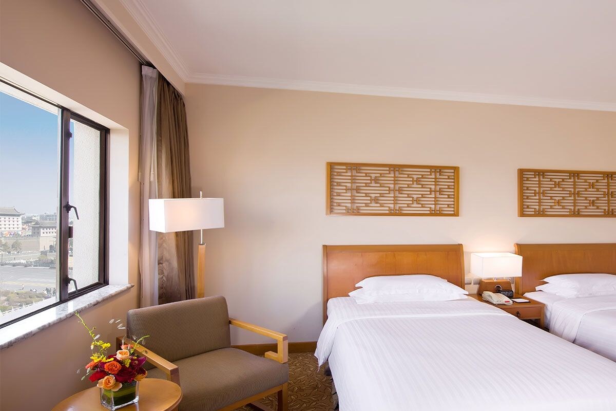 A hotel room with two beds, a chair, a small table with a vase of flowers, and a view of the city through the window.