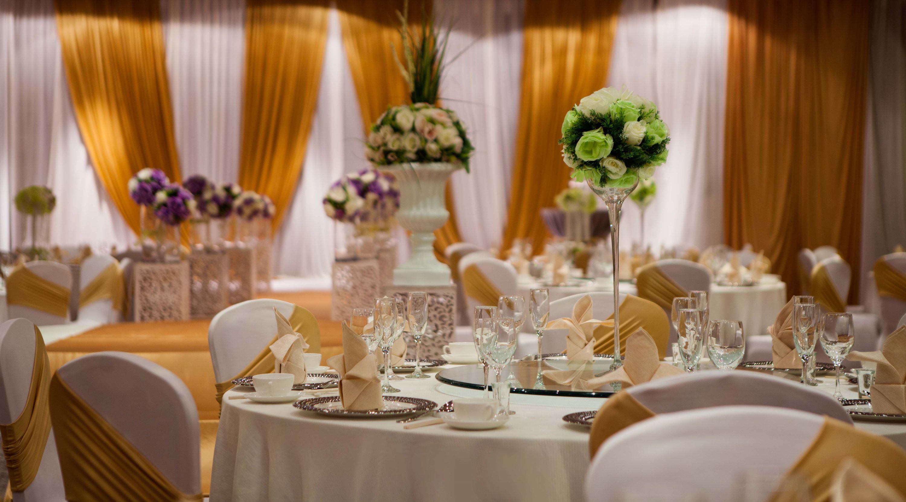 Un salón de banquetes con mesas redondas preparadas para un evento formal, con manteles blancos, fundas doradas en las sillas y centros de mesa florales.